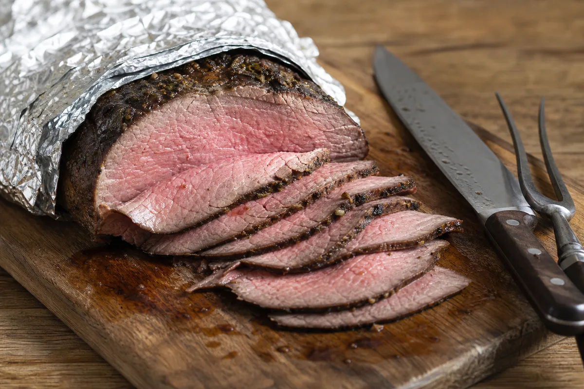 Tranches fines de rôti de bœuf au repos sur une planche en bois, recouvertes sans serrer d&rsquo;une feuille d&rsquo;aluminium